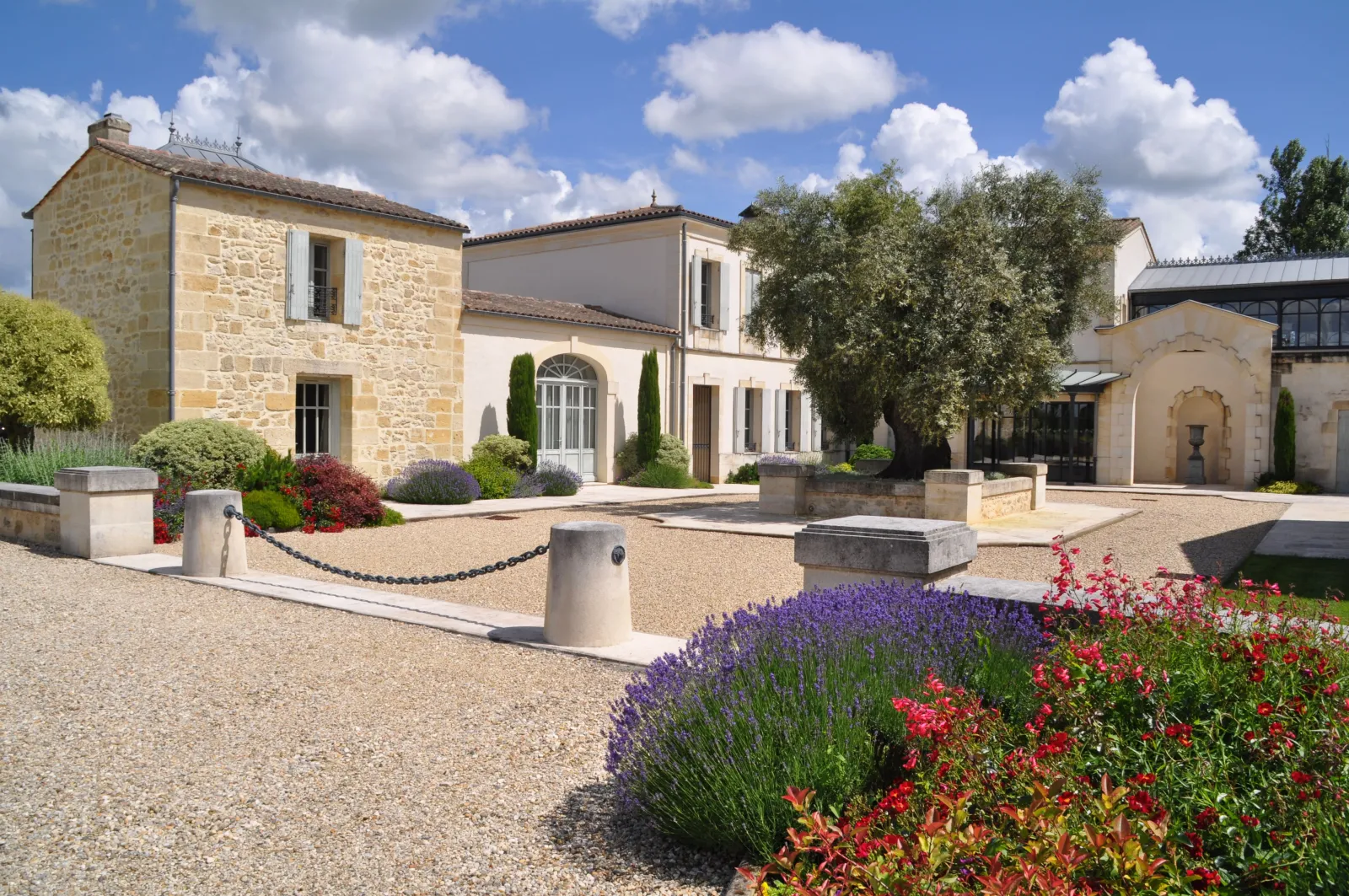 Elegant courtyard of Château La Rose Perrière near Bordeaux, featuring white stone buildings, lavender and red flowers under a partly cloudy blue sky.