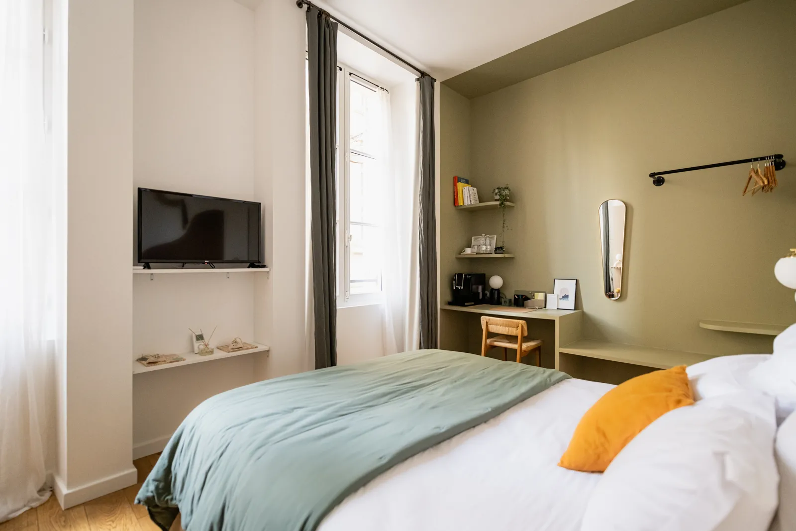 Modern bedroom at La Maison Fernand in Bordeaux, featuring a white and blue bed, wall-mounted TV, wooden desk, floating shelves, round mirror, gray curtains, and sage green walls.