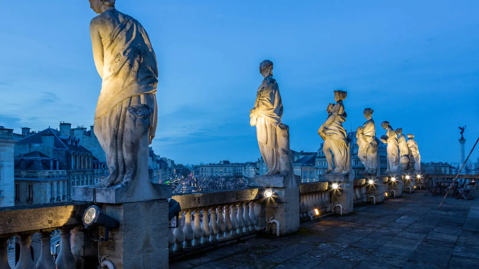 Illuminated statues on the roof of the Grand Theatre of Bordeaux at dusk, overlooking the historic cityscape below.