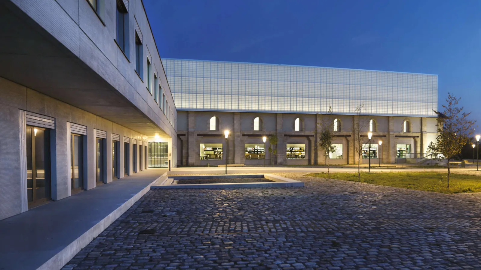 Modern Archives Bordeaux Métropole building photographed at dusk, featuring white brick facade, large illuminated windows and paved courtyard
