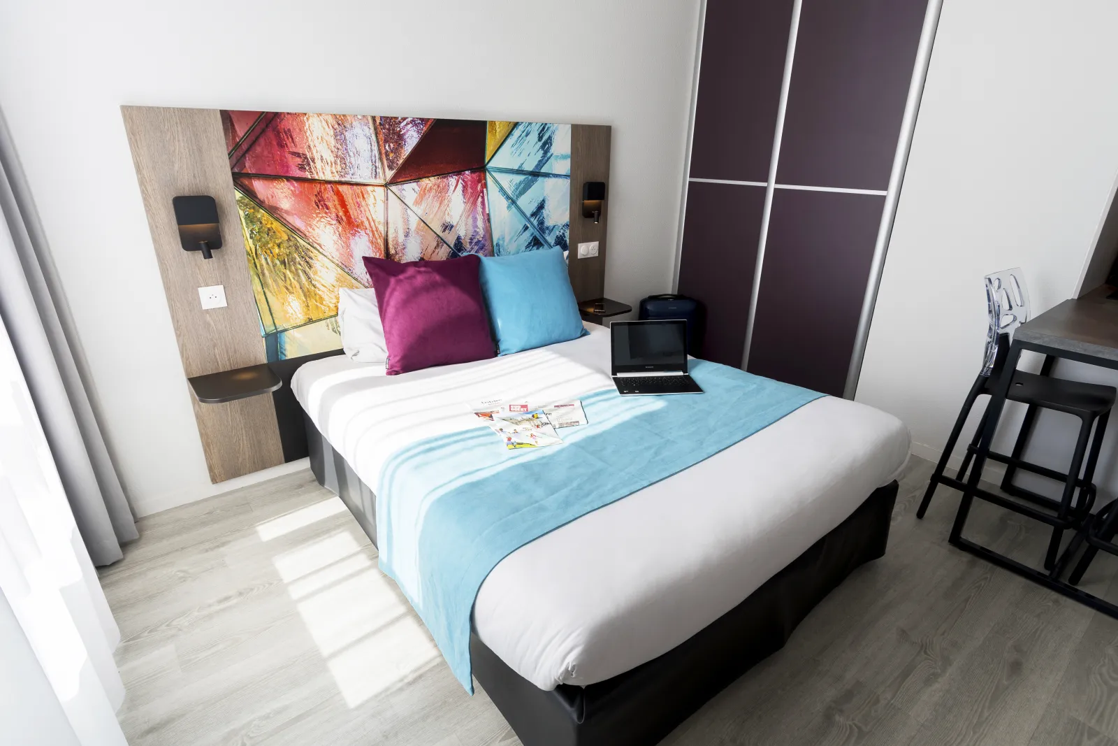Modern hotel apartment bedroom in Bordeaux featuring a double bed with light blue linens, magenta and turquoise pillows, wooden headboard with colorful geometric artwork, purple wardrobe, black desk, and gray curtained windows.