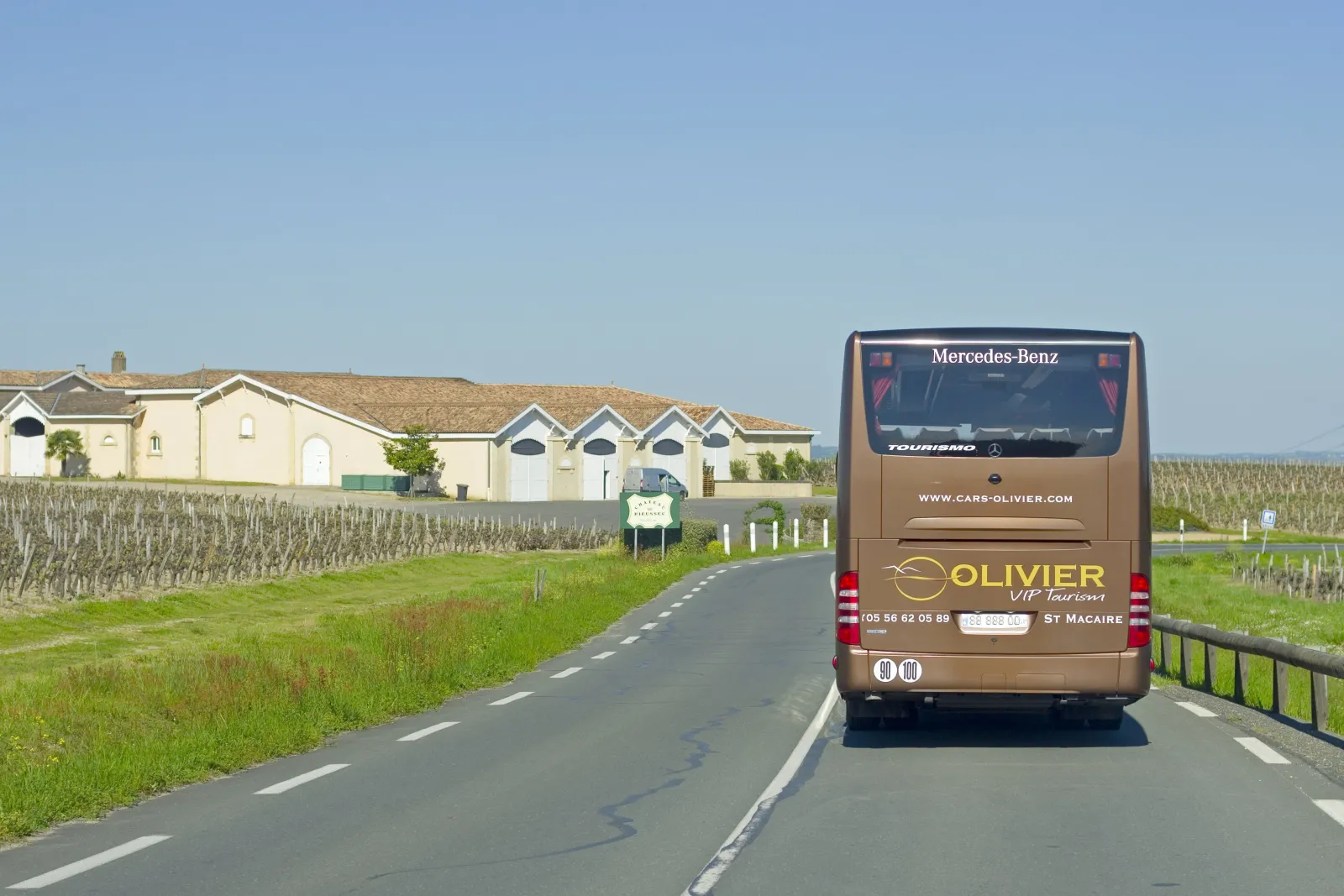 A Mercedes-Benz Tourismo coach from Cars Olivier drives on a rural road lined with vineyards in Gironde, with typical wine-producing buildings in the background.
