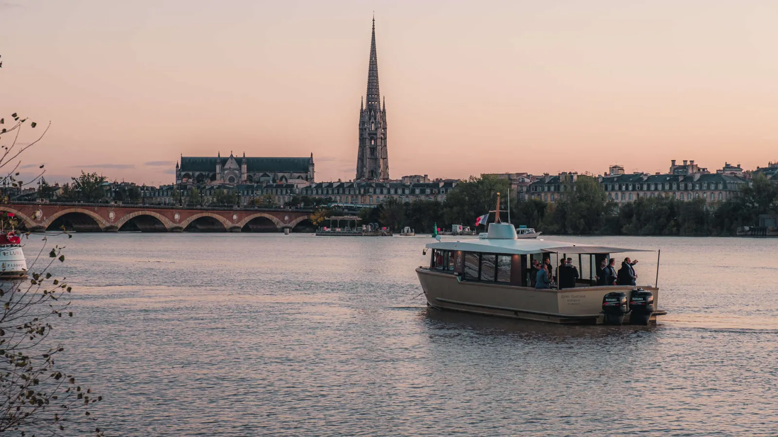 Sunset view of Bordeaux featuring the Saint-Michel Spire in the background, the Stone Bridge spanning the Garonne River, and a tour boat in the foreground.