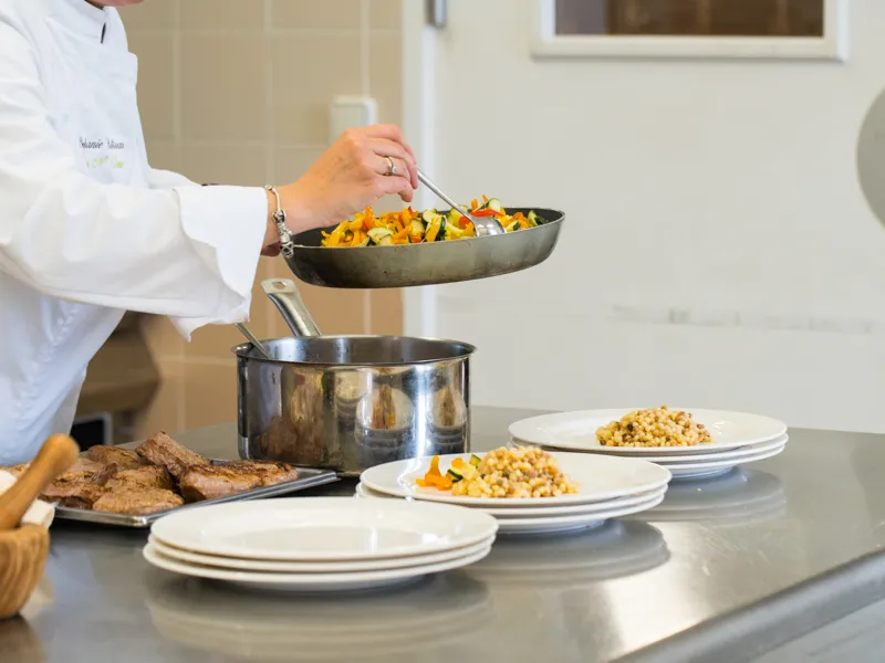 A chef in white jacket transfers sautéed vegetables from a pan into a cooking pot during a culinary workshop at Château Kirwan in Bordeaux. Plates with corn and vegetables are arranged on the work surface.