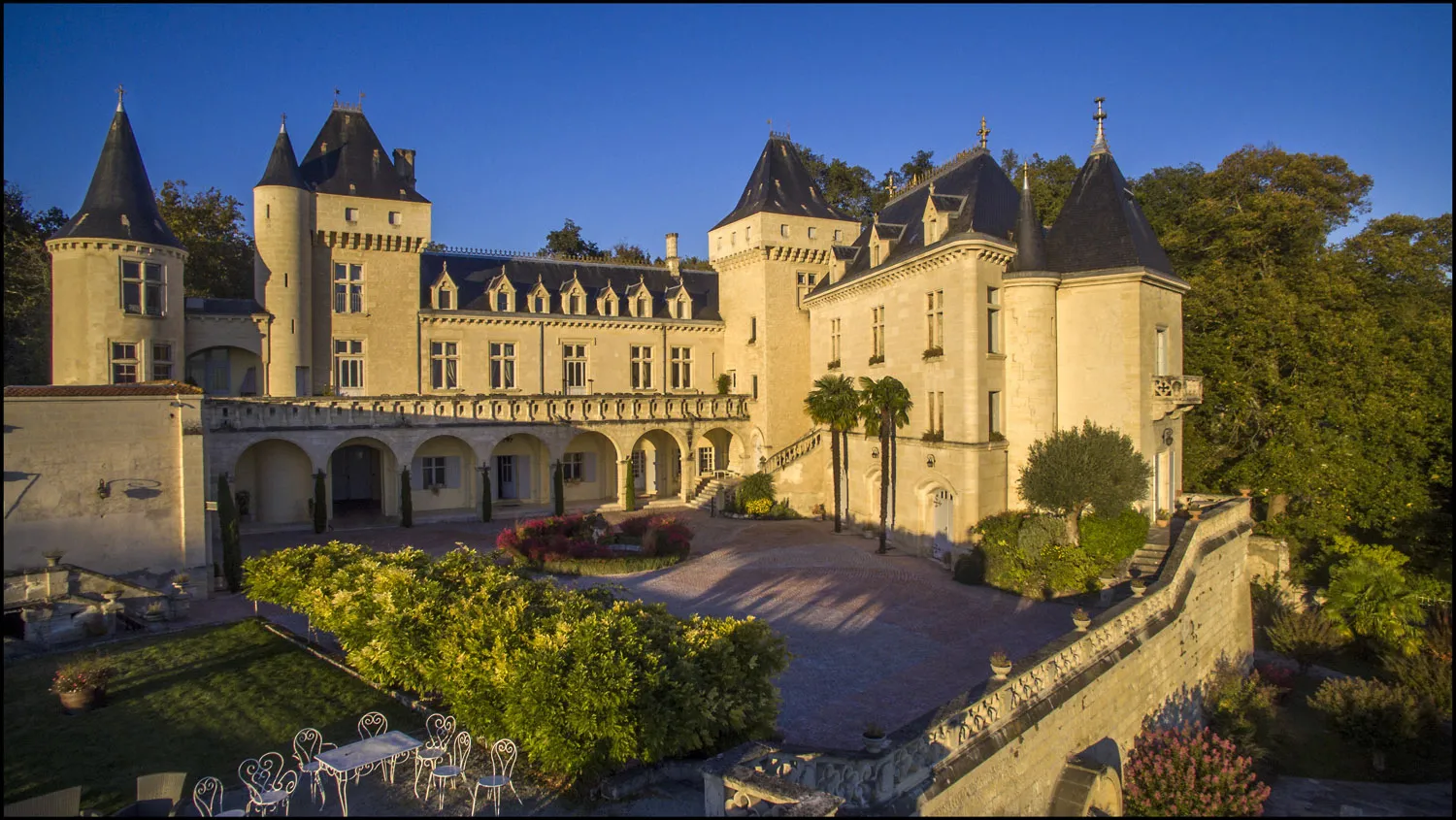 Sunny view of Château de La Rivière in Bordeaux, featuring pointed towers, pale yellow façade, paved courtyard with flowers and stone balustrade.