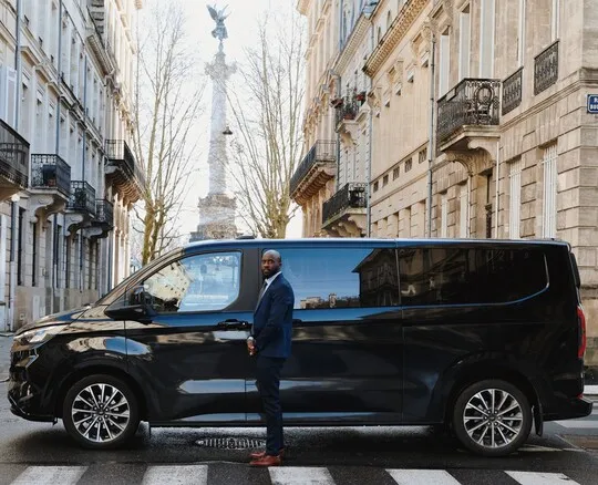 A man in a navy blue suit stands beside a luxurious black minivan on a paved Bordeaux street, with Haussmann buildings and the Monument to the Girondins visible in the background.