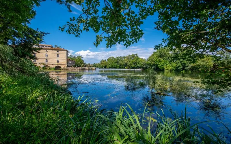 Porchères Mill in Bordeaux, a historic stone building beside a calm river surrounded by lush green vegetation and trees.