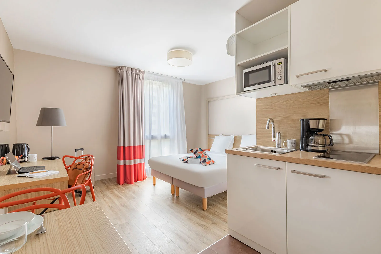 Modern studio apartment at Appart City Confort Bordeaux Chartrons featuring equipped kitchenette, queen-size bed, work area, orange chair and red and white curtains