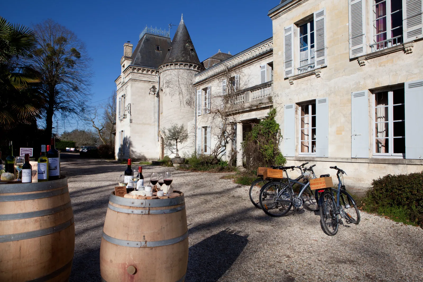 Inner courtyard of Château des Ateliers in Bordeaux featuring a wine barrel, rental bicycles, displayed wine bottles and glasses, white stone facade and conical tower in the background