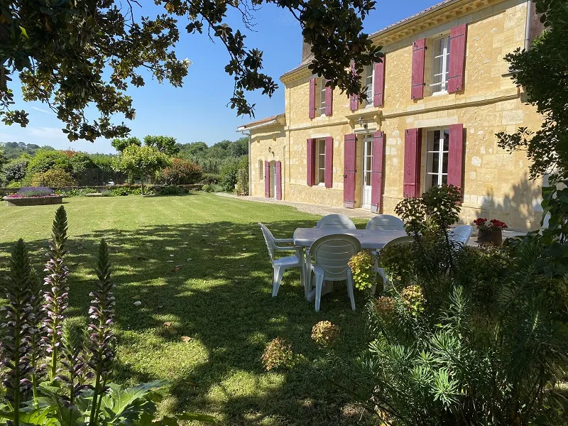 La Gontrie Castle in Bordeaux featuring a yellow stone facade with pink shutters, green lawn with white chairs and flowering gardens