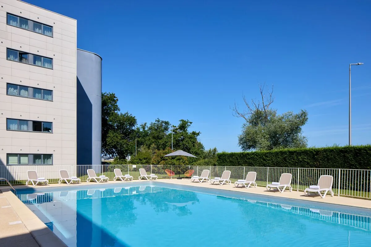 Outdoor swimming pool at Zenitude Bordeaux Airport hotel featuring white loungers, modern building in background, and clear blue sky.