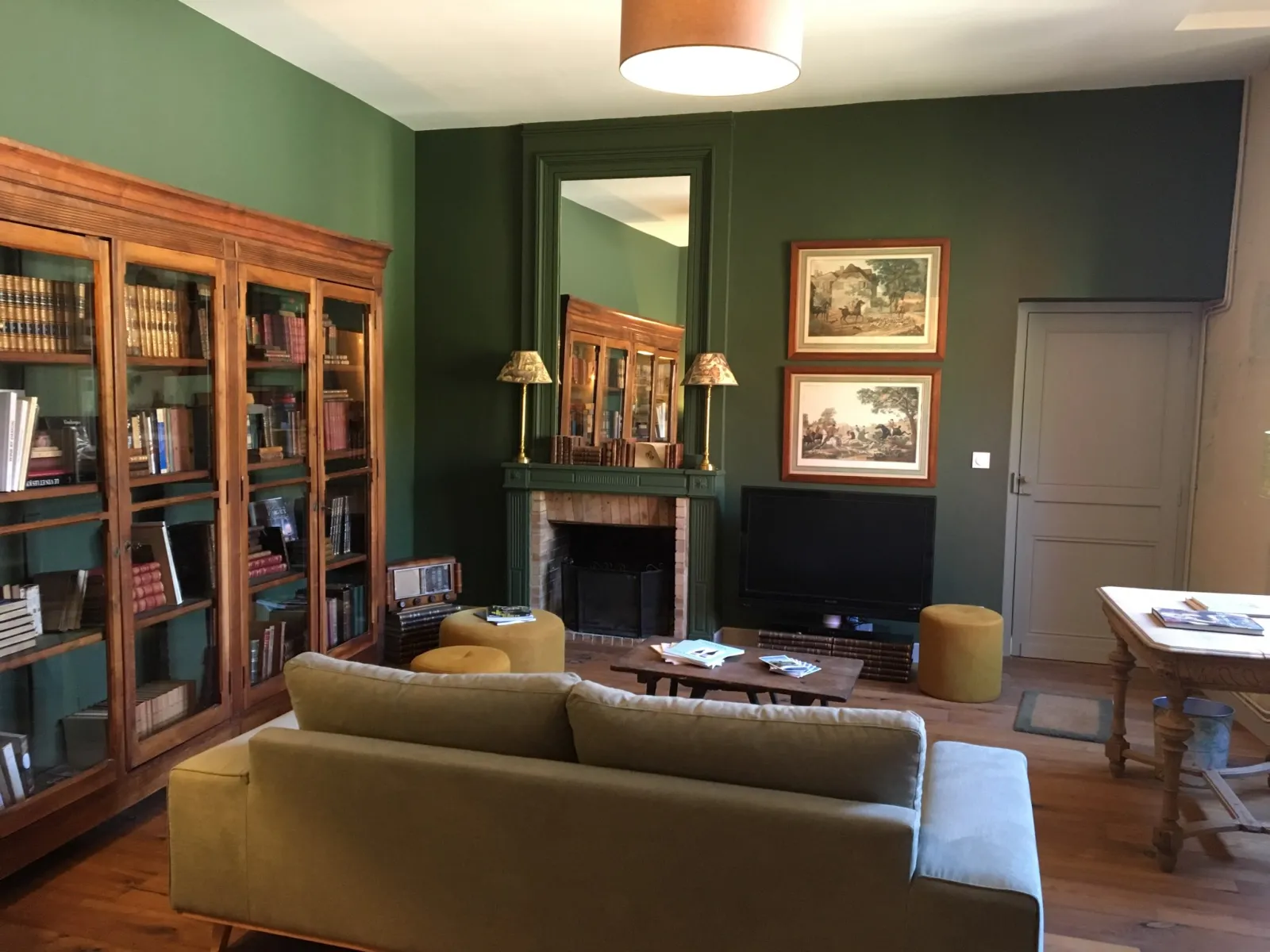 Elegant living room at Château Sigalas Rabaud in Bordeaux featuring forest green walls, wooden glass-fronted bookcase, fireplace, beige sofa, mirror and framed vintage engravings