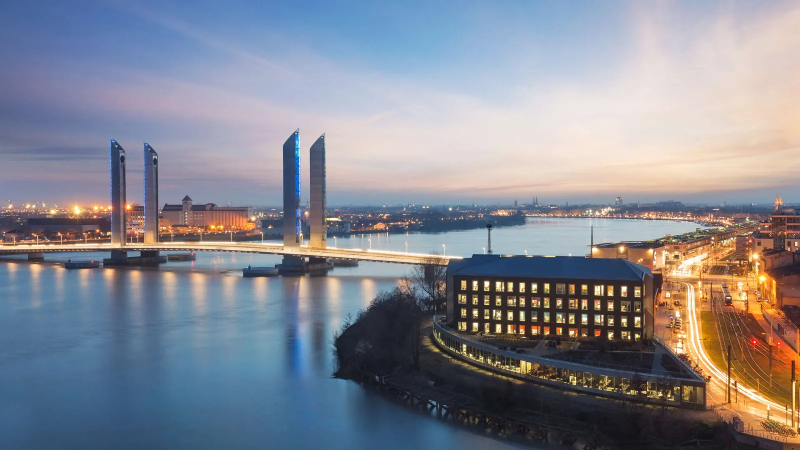 Twilight view of Bordeaux featuring the illuminated Jacques Chaban Delmas Bridge with its distinctive blue twin towers reflected in the Garonne river, surrounded by historic and modern buildings.