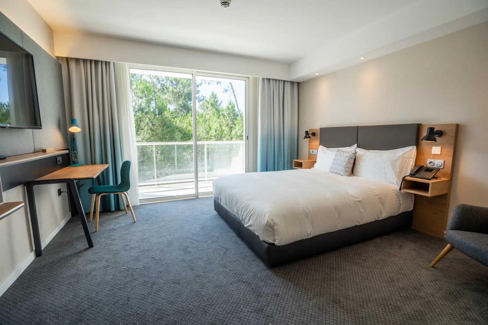 Modern hotel room at Holiday Inn Bordeaux Mérignac featuring a queen-size bed, work desk, wall-mounted TV, and glass balcony overlooking trees