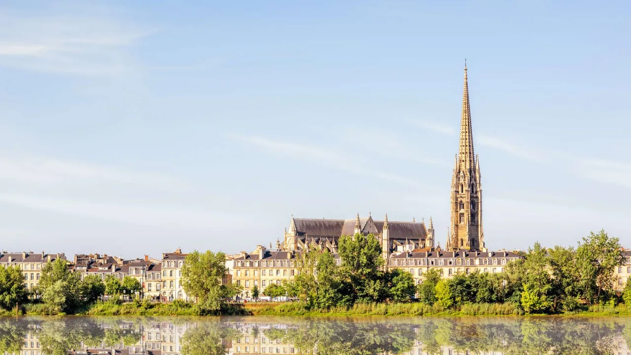 Panoramic view of the Basilica of Saint-Michel in Bordeaux with its Gothic spire towering over the urban landscape, surrounded by Haussmann-style buildings and reflected in the Garonne river waters.