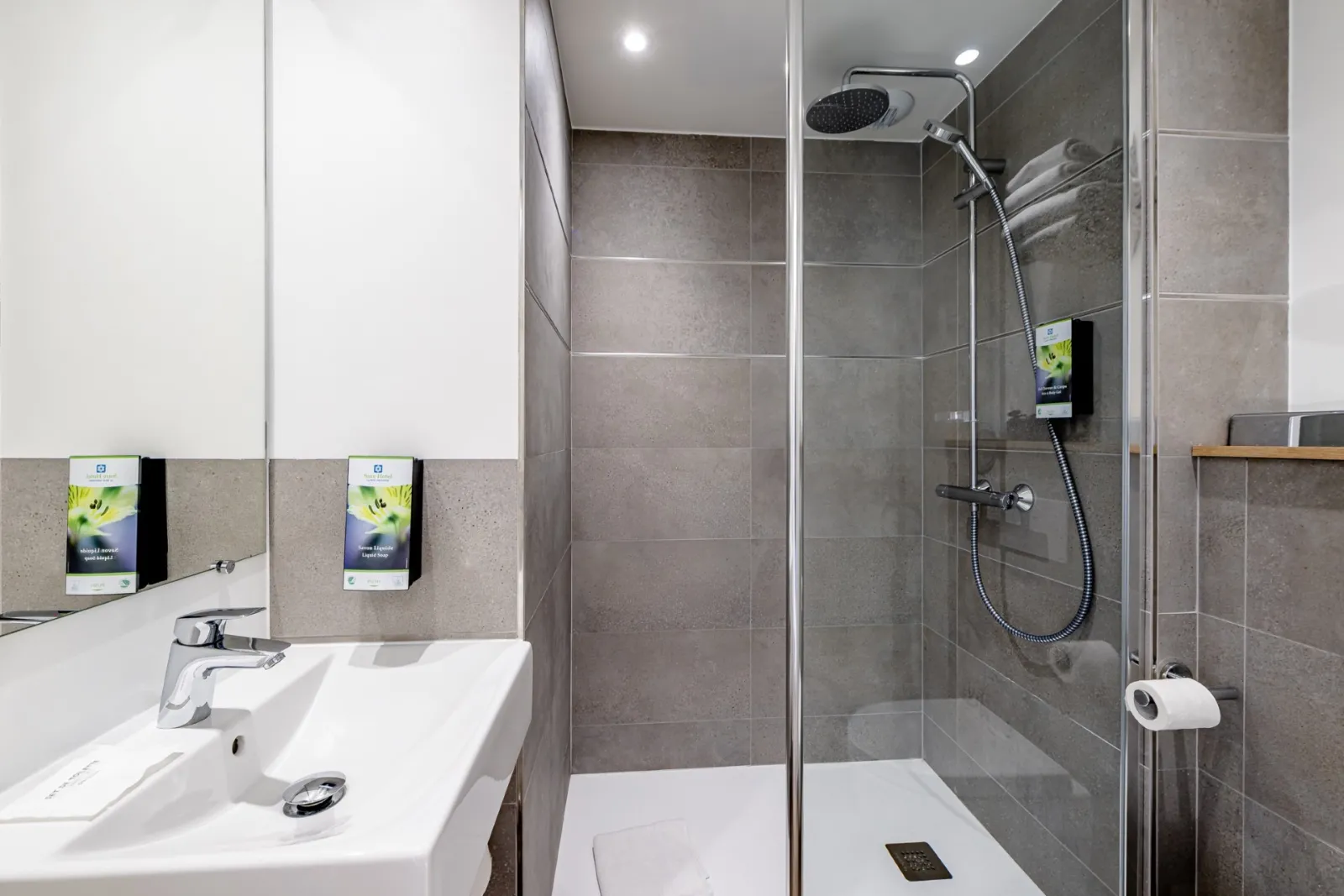 Modern bathroom featuring a white sink, mirror, soap dispensers, and walk-in shower with glass partition and gray tiling at Sure Hotel by Best Western Bordeaux Lac