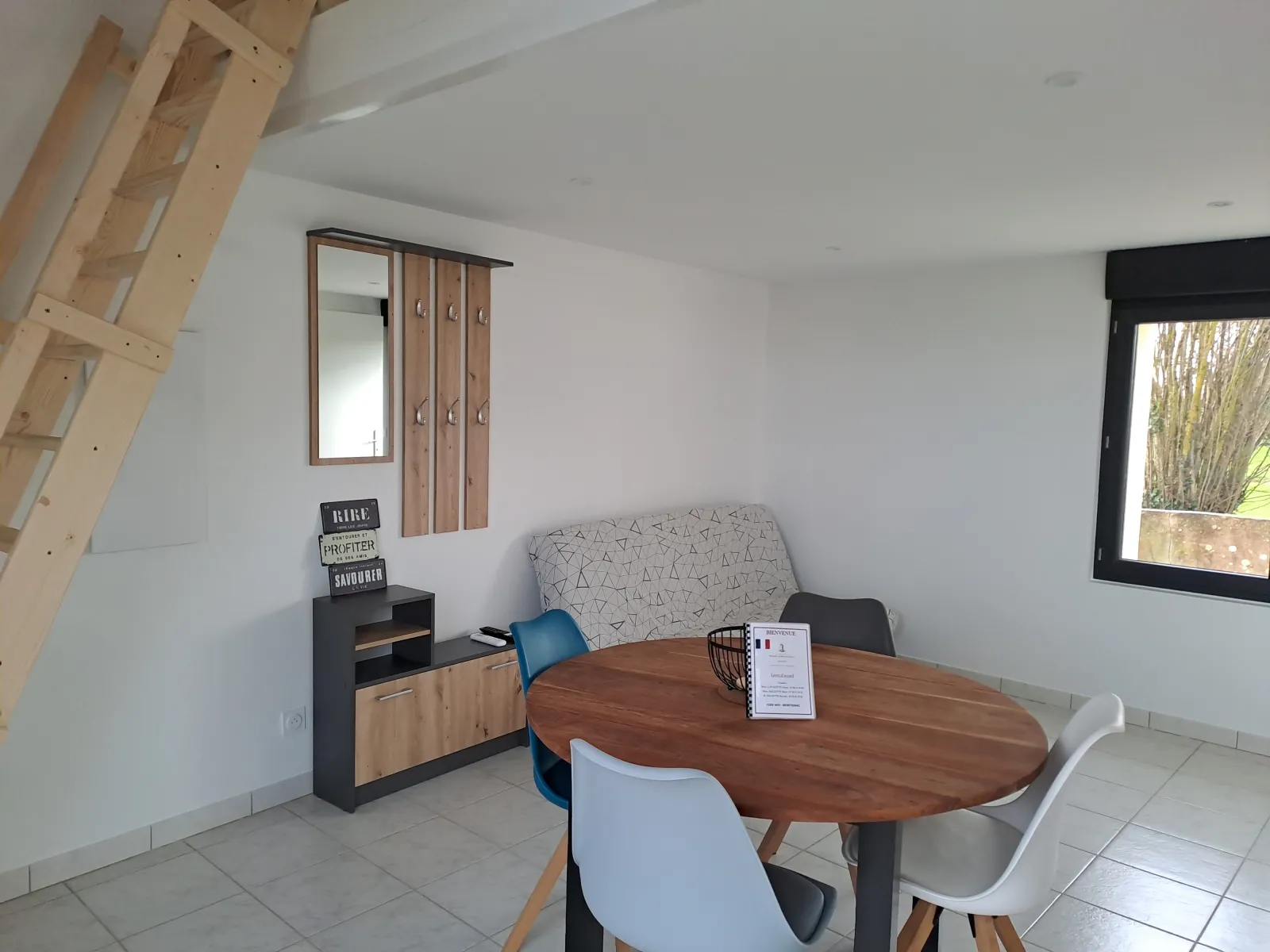 Modern living area of Anthocyane cottage featuring a round wooden table, colored chairs, gray sofa, black and wood storage furniture, wall mirror, light wooden staircase and window overlooking greenery