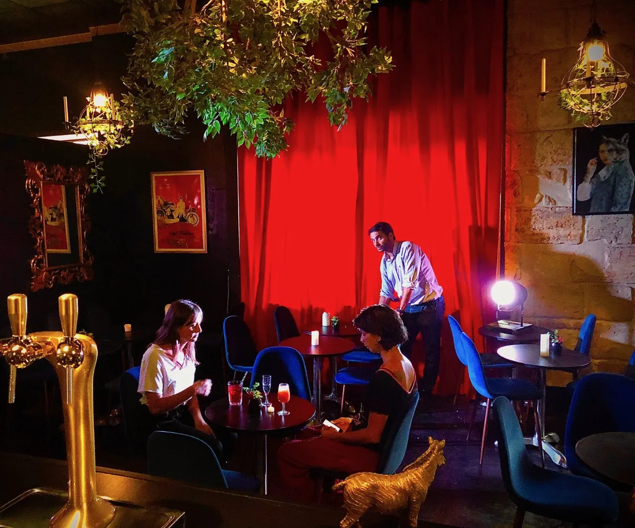Interior of Théâtre des Chartrons in Bordeaux featuring three people in an elegant bar with red walls, blue furniture, sophisticated lighting and plant decoration.
