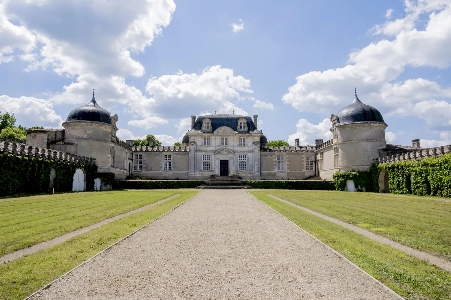 Front view of Château de Malle in Bordeaux, featuring two round towers with black domes, white facades, and symmetrical green pathways under a clear sky.