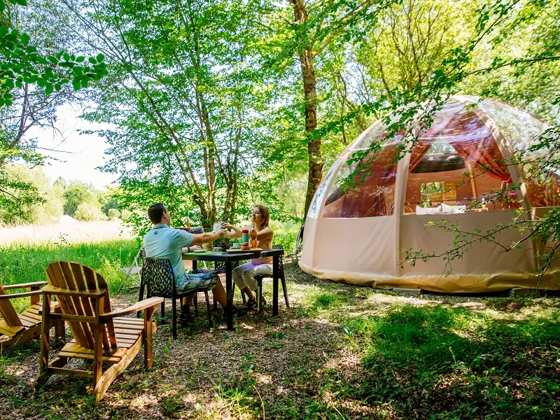 Two people dining at a wooden table in front of a transparent dome-shaped glamping accommodation, surrounded by Adirondack chairs and lush green trees at EcÔtelia Domain in Bordeaux.