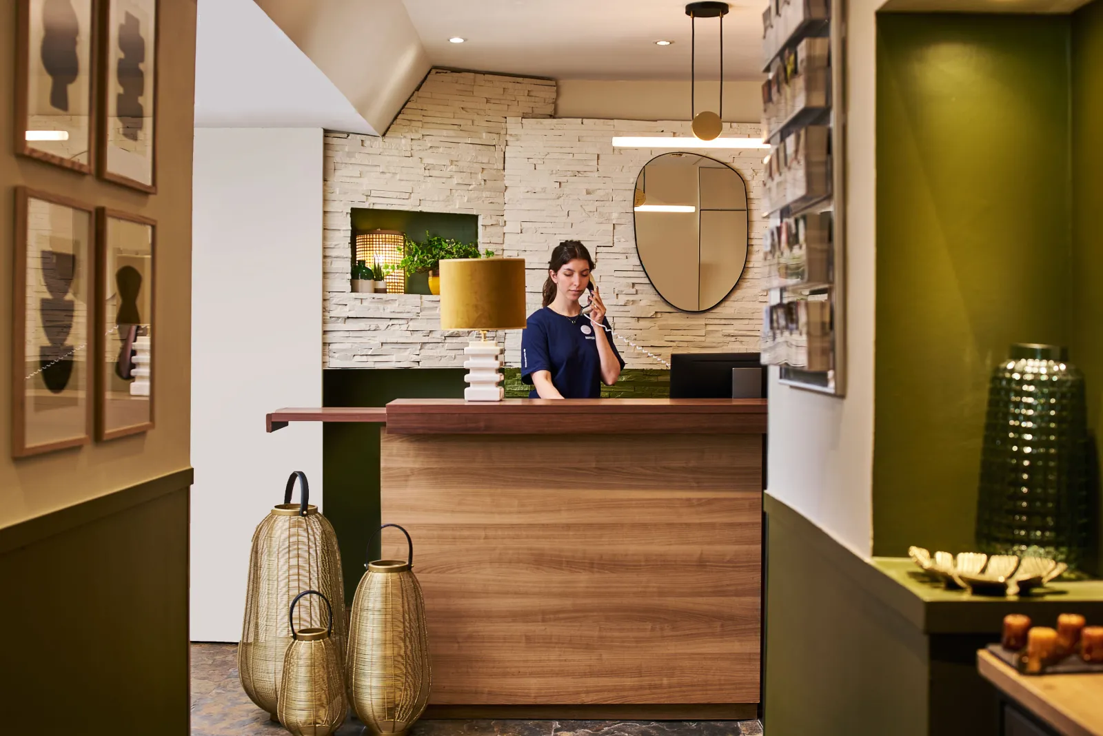 Modern hotel reception desk with staff member, wooden counter, white stone walls, oval mirror, green and natural decor elements, and woven baskets in the foreground.