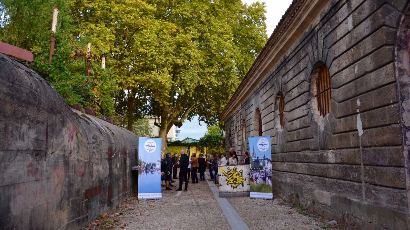 Narrow stone-walled passage in Bordeaux with Les Vivres de l'Art event banners, visitors in the background, and golden-leafed trees overhead.