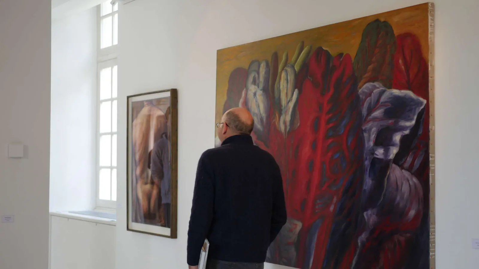 A visitor viewing a large abstract painting with red and gold tones in a contemporary art gallery, with additional artworks visible on the walls and natural light from a window on the left.