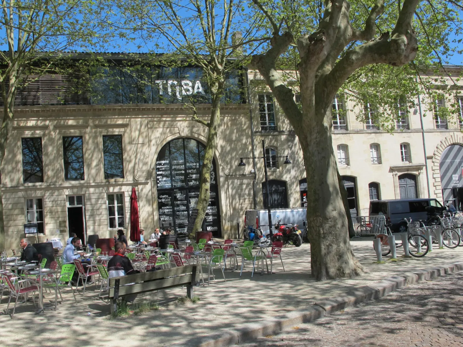 Outdoor café terrace with colorful tables and chairs in front of the Abbatiale Sainte-Croix in Bordeaux, shaded by large trees on a sunny day