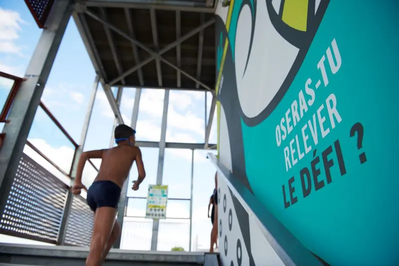 A child runs on a trampoline in a covered aquatic center with a turquoise sign displaying French text