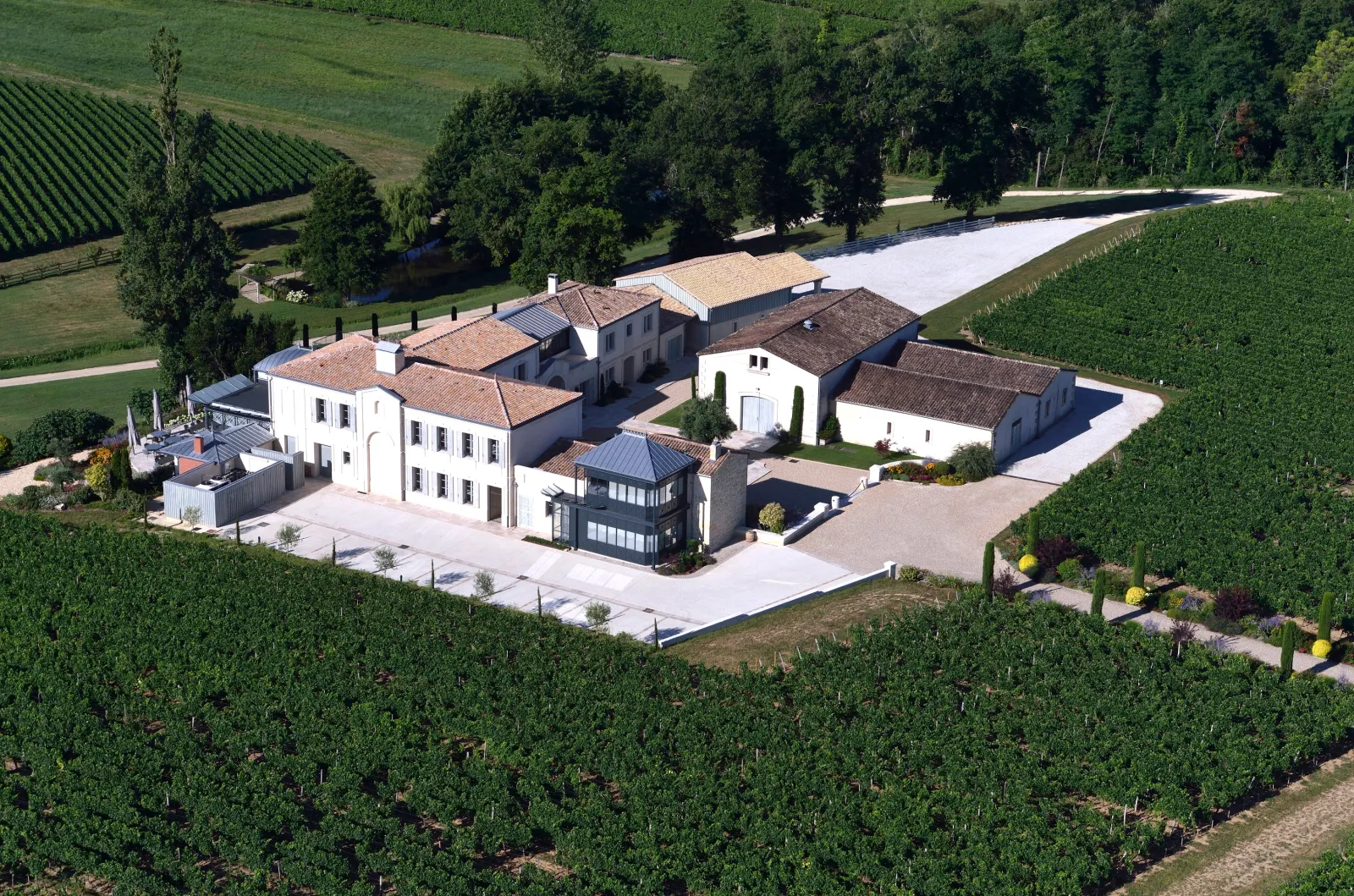 Aerial view of Château La Rose Perrière near Bordeaux, featuring modern white renovated buildings surrounded by vineyards and a river.