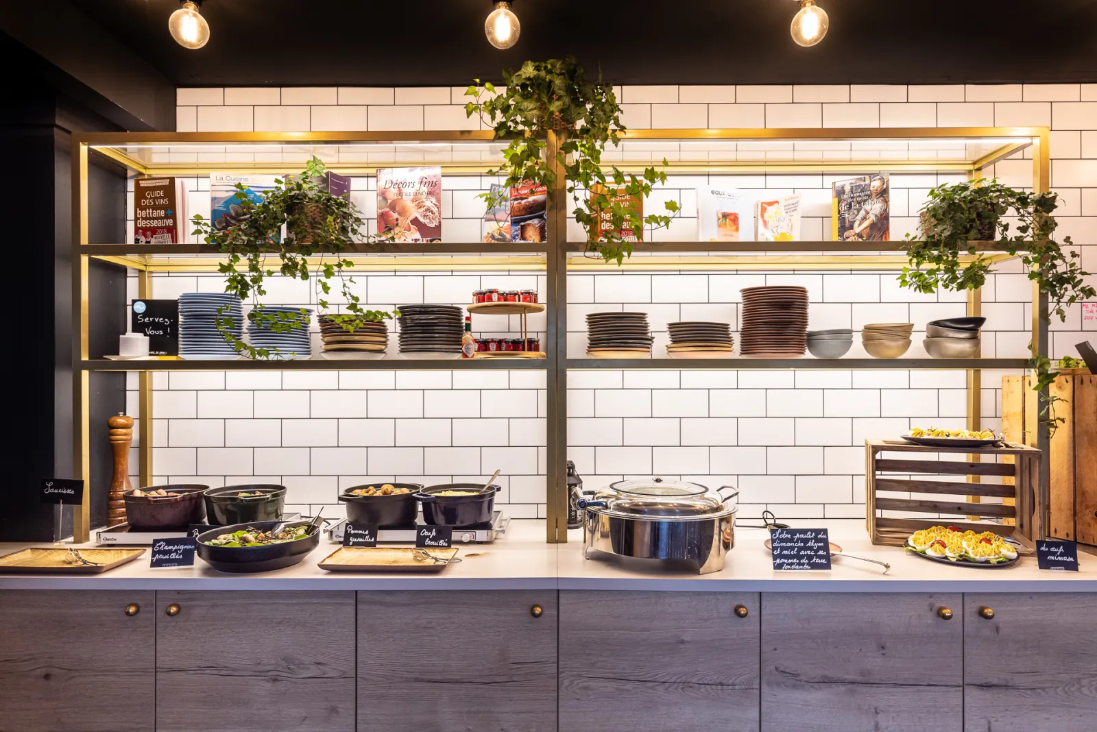 Restaurant window display showcasing cooked dishes in black pans, stacked plates, and kitchen accessories on gold shelves with white subway tiles and living plant decoration.