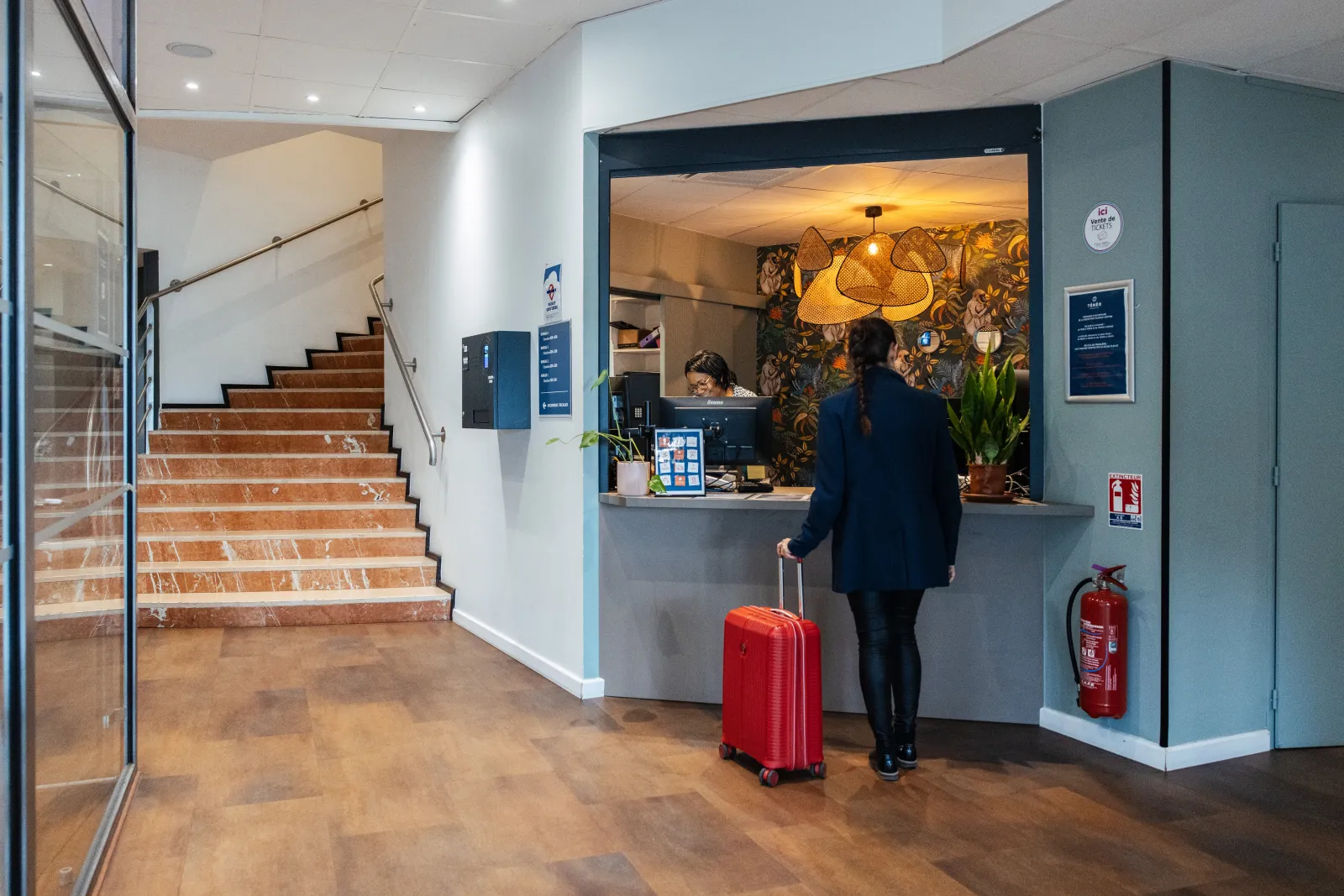 Modern lobby of Ténéo Apparthotel Talence Centre in Bordeaux, featuring reception desk, marble staircase, guest with red suitcase, colorful wall art and designer lighting.