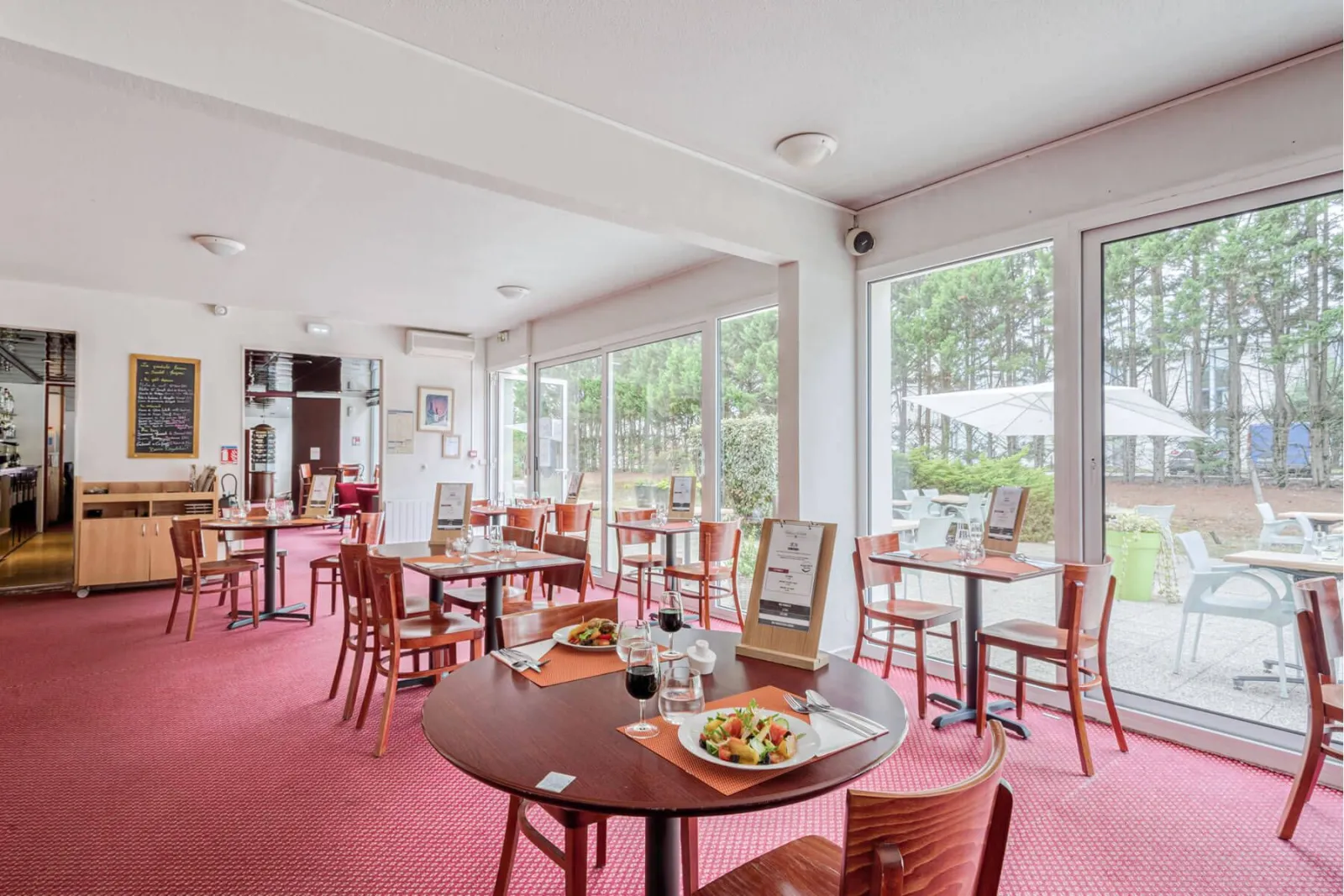 Modern restaurant dining room at Soretel Bordeaux Mérignac featuring round and square tables, wooden chairs, pink carpet, and large glass doors opening onto an outdoor terrace with white umbrellas and forest views.