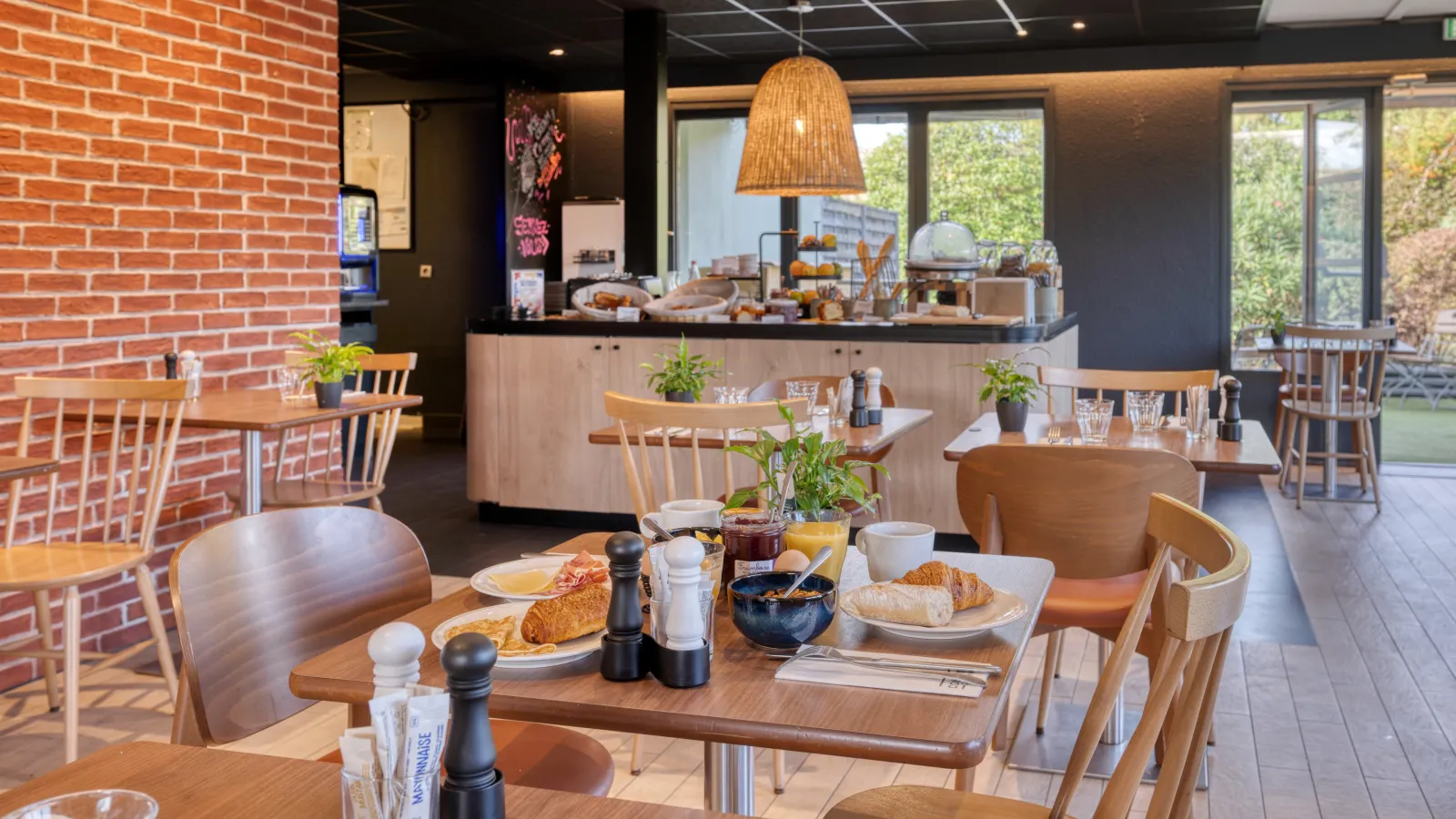 Modern breakfast room at a Campanile hotel featuring wooden tables, Scandinavian chairs, red brick wall, service counter with pastries and beverages, and large glass doors overlooking the garden.