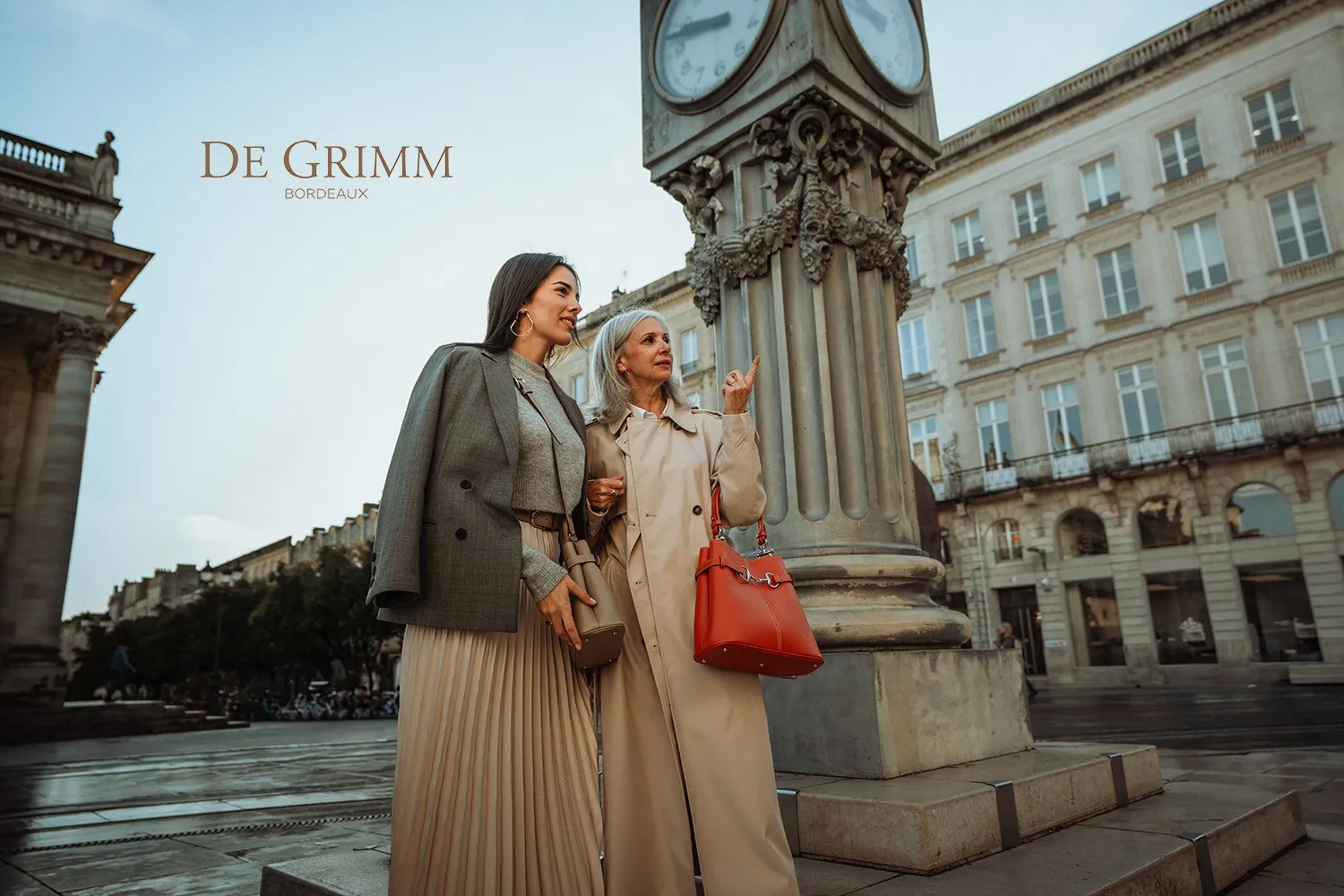Two stylish women strolling past an ornamental clock column in Bordeaux, with Haussmann-style buildings in the background