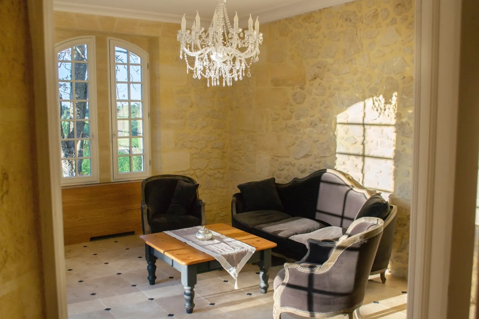 Elegant living room with yellow damask walls, white crystal chandelier, grey and black armchairs, wooden coffee table, large windows overlooking gardens.