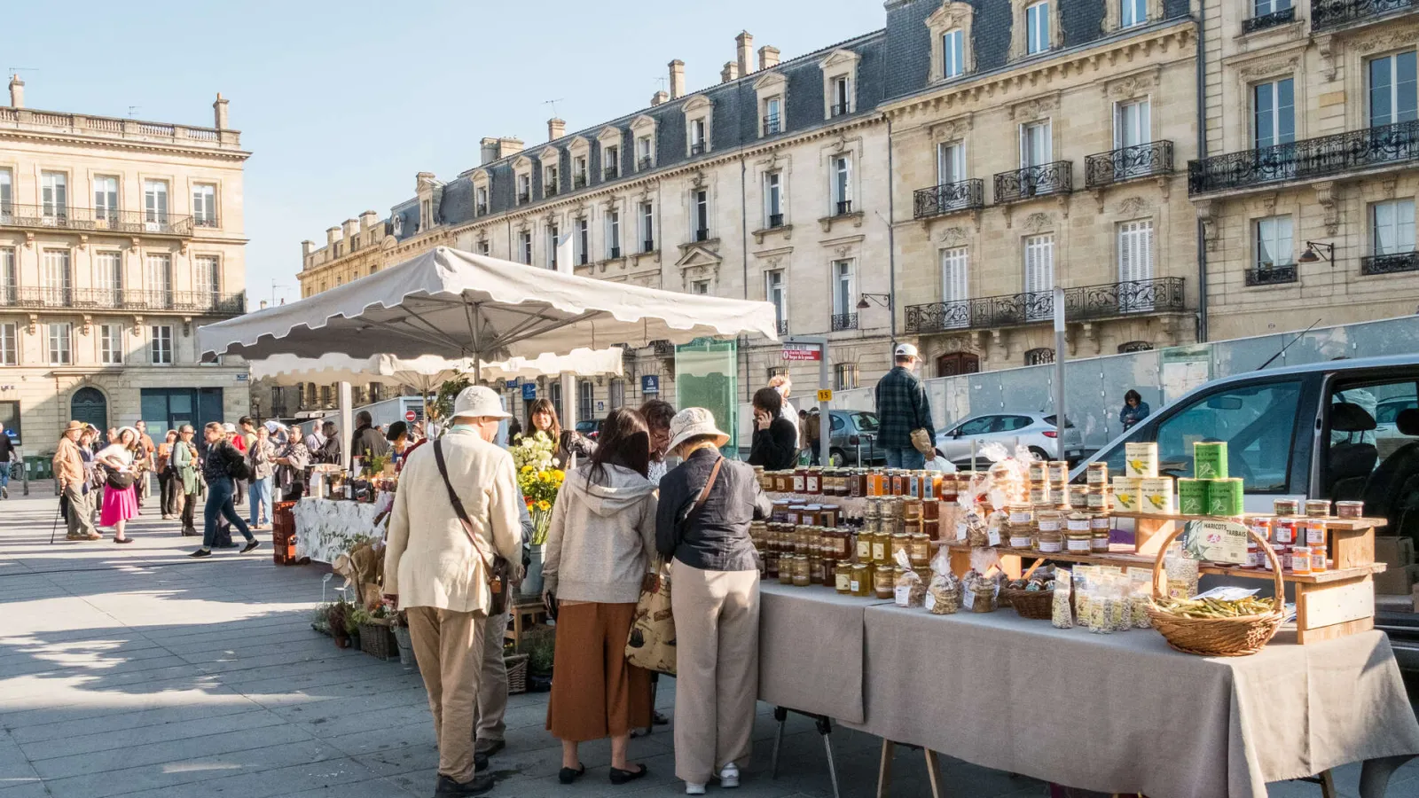 Outdoor market in Bordeaux featuring local vendors selling products under white umbrellas, with Haussmann-style architecture visible in the background.