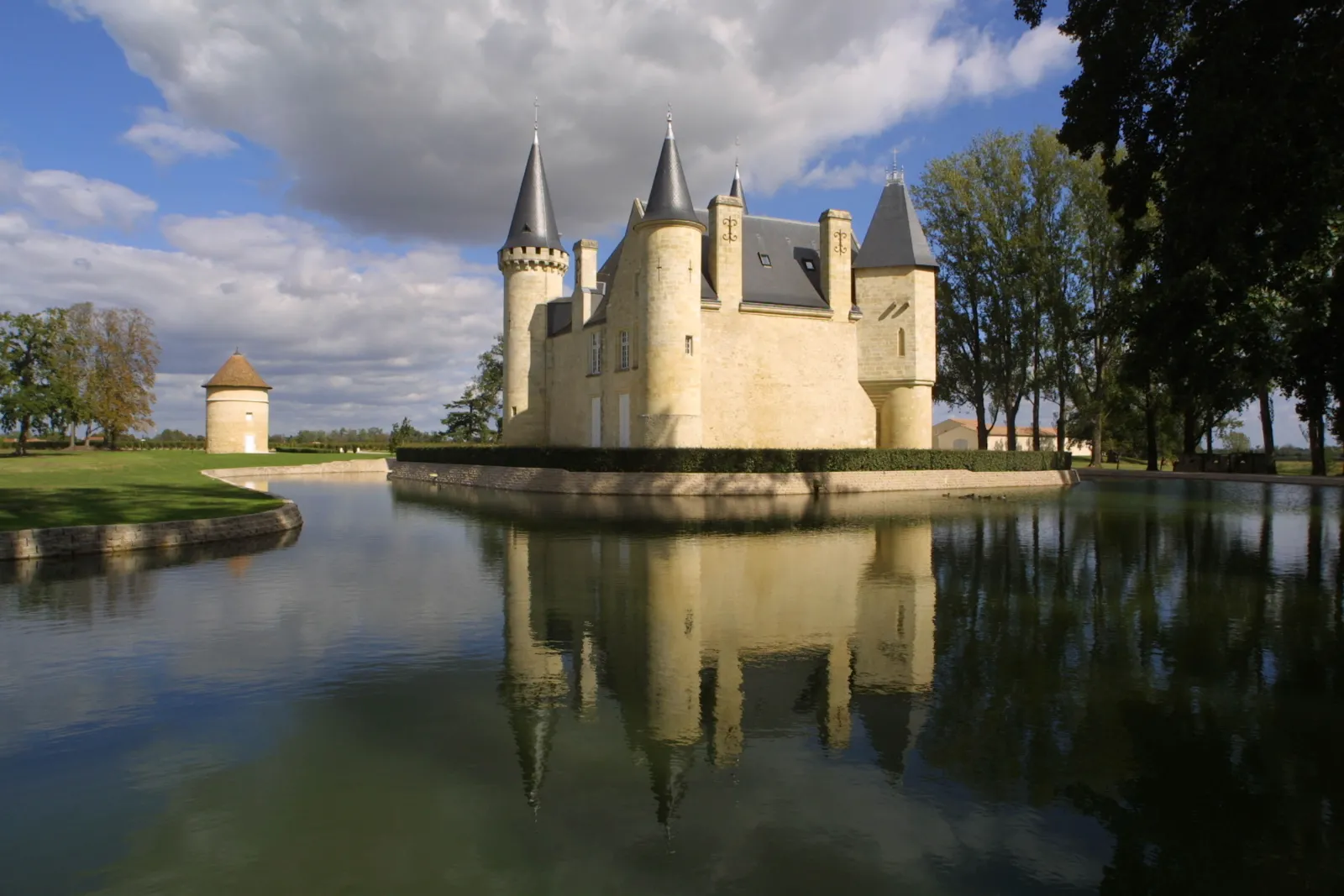 Château d'Agassac in Bordeaux, a medieval fortress with pointed towers surrounded by a water-filled moat, reflected in the basin, with a small watchtower nearby and trees around it.
