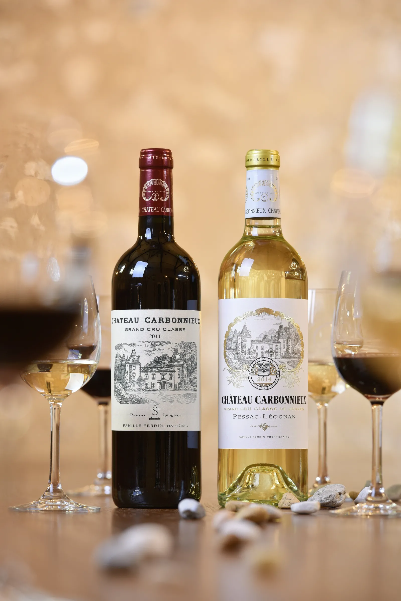 Two bottles of Château Carbonnieux, a red wine and a white wine from Bordeaux, surrounded by filled glasses and grapes on a golden surface with a blurred background.