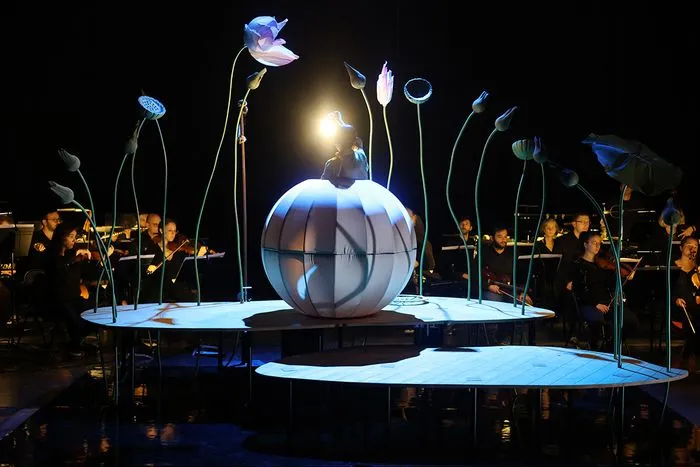 Nighttime performance scene in Bordeaux featuring a large illuminated sphere at the center surrounded by stylized flower stems and musicians in black on both sides for the show 'Momo, the Peach Child'.