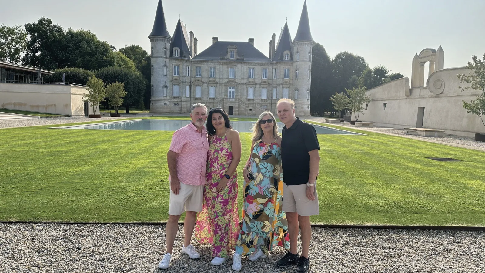 Four tourists posing in front of a castle with pointed towers, a rectangular pool and green lawn in Aquitaine