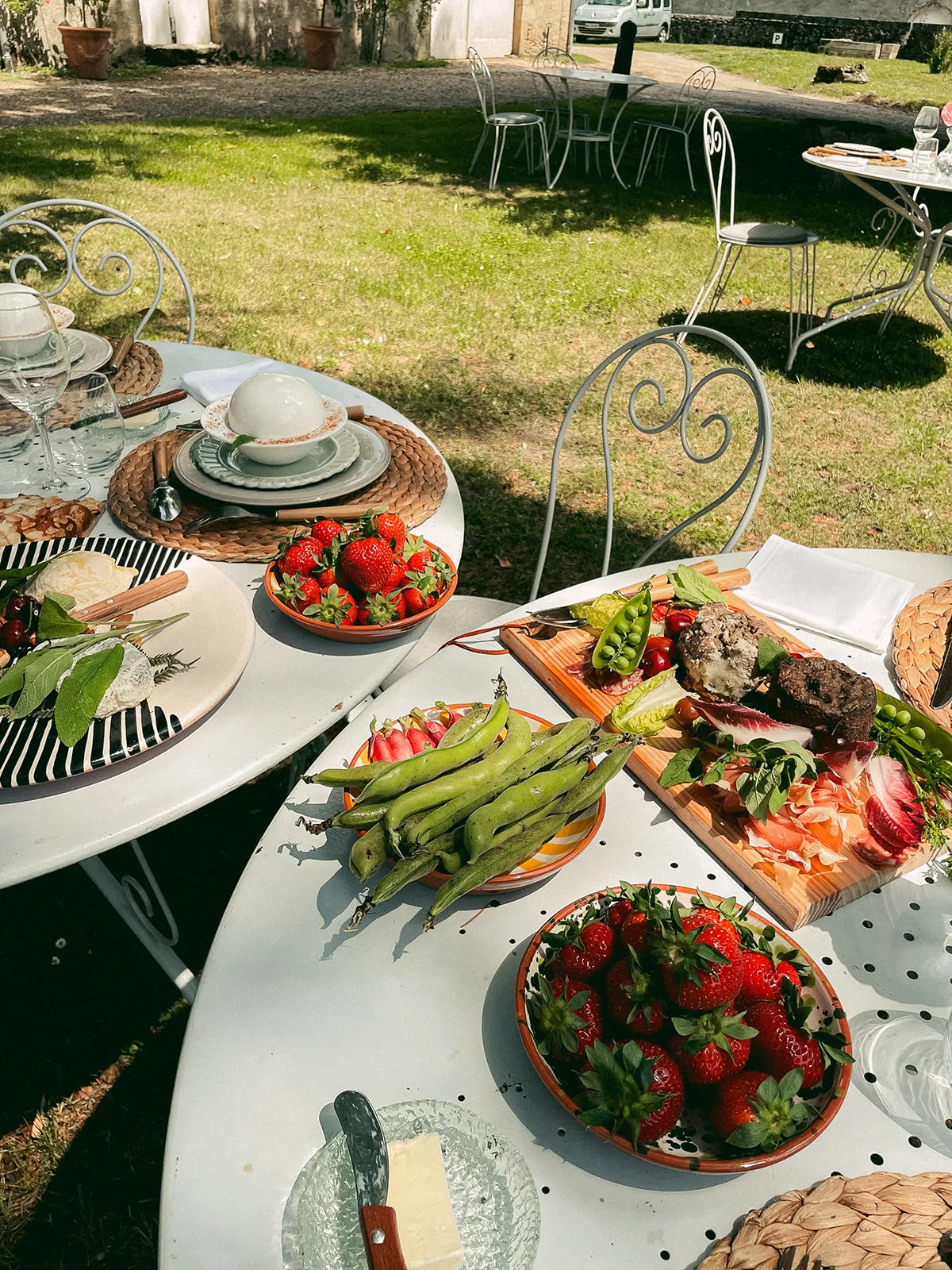Outdoor dining table at Château de Cérons in Bordeaux, decorated with fresh strawberries, asparagus, cold cuts, bread and cheese, with wrought iron chairs and set tables in a sunny garden.