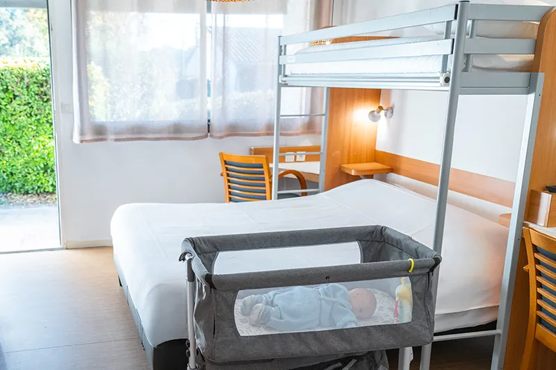 Hotel room featuring a double bed, bunk bed, portable baby crib, and light wooden furniture with glass doors overlooking a garden