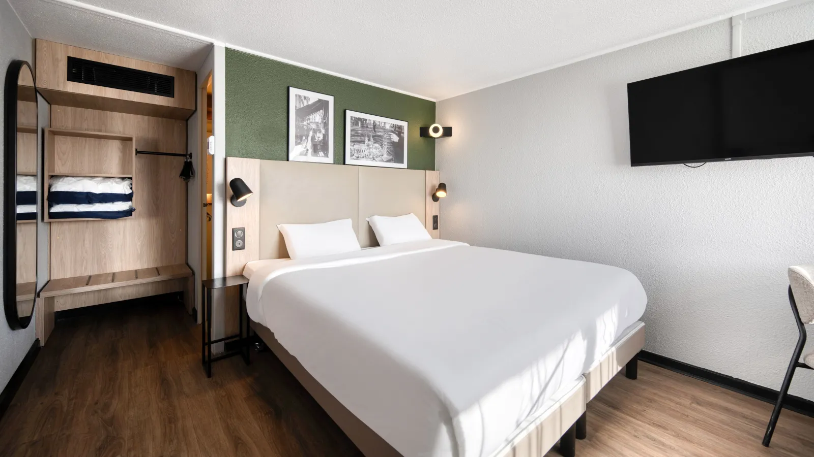 Modern hotel room featuring a double bed with white linens, beige headboard, green accent wall, wall-mounted TV, light wood wardrobe and bunk beds in the background