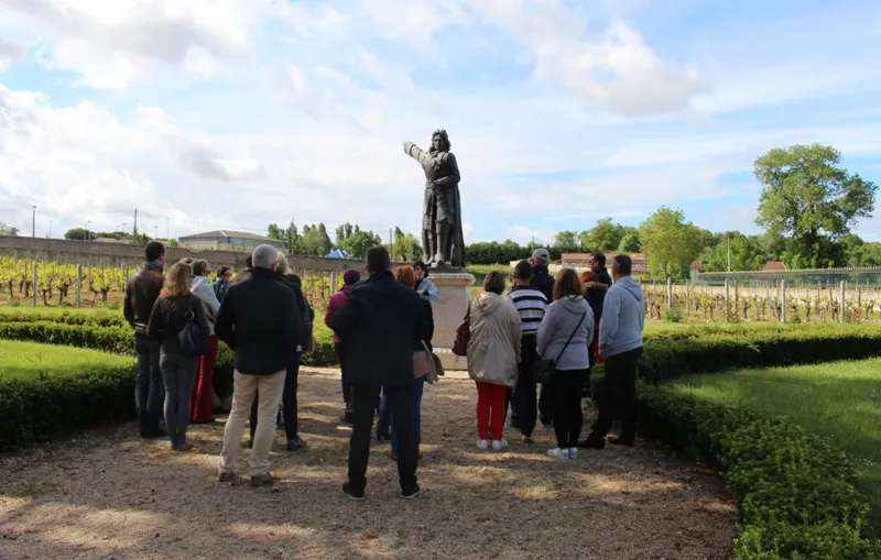 Group of visitors gathered around a bronze statue in the vineyards of Château Marquis de Vauban near Bordeaux, with rows of grapevines and cloudy blue sky in the background.
