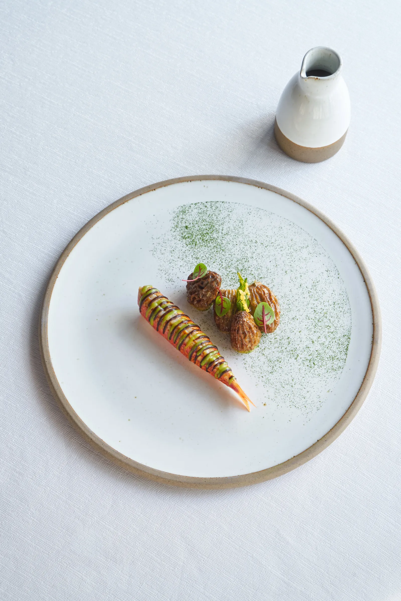 Gourmet plated dish featuring a striped carrot, two meat-based preparations, and microgreens, with a white and beige sauce pot