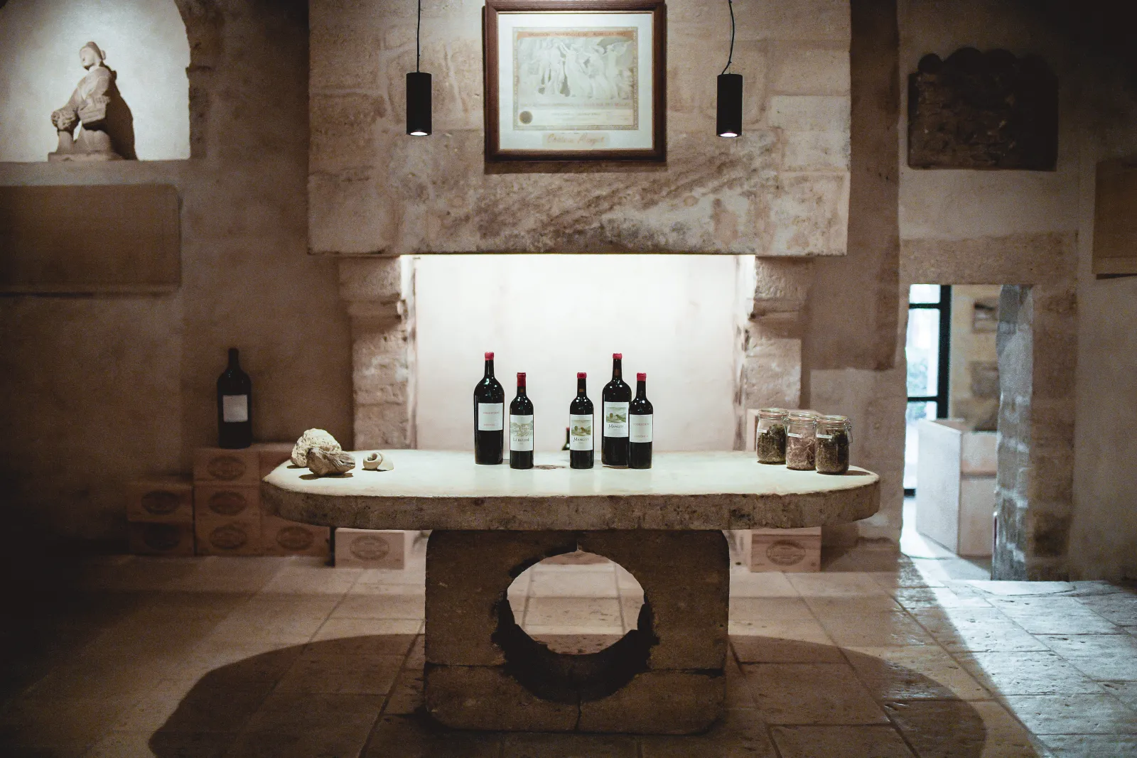 Interior of Château Mangot in Bordeaux: a round stone table displaying red wine bottles and glass jars, in a stone cellar with aged walls, lit by black pendant lights.