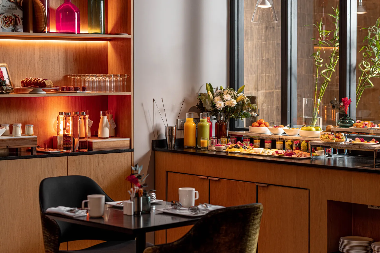 Elegant breakfast area at Hôtel Villas Foch in Bordeaux, featuring modern furniture, warm wooden shelving with ambient lighting, pastry display bar with fresh juices, flowers, and large windows.