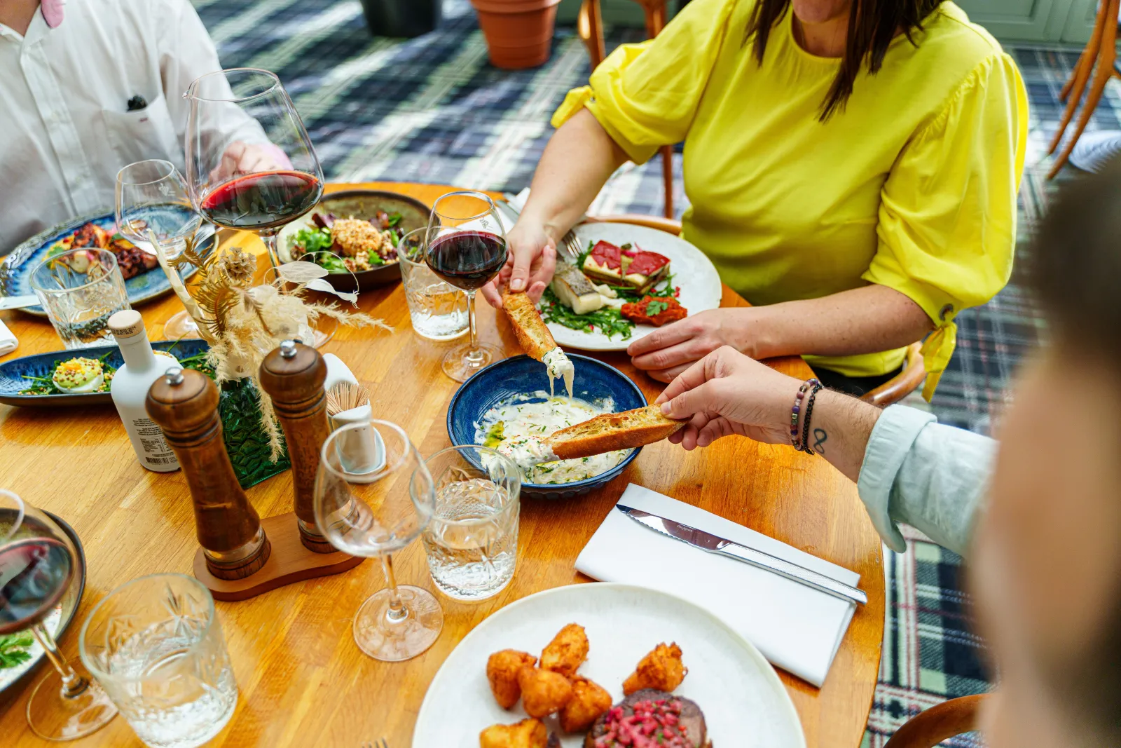 Dining scene at C Restaurant in Bordeaux: a woman in a yellow shirt serves toasted bread with a dip bowl, surrounded by red wines, various dishes, and guests enjoying a gastronomic meal together.