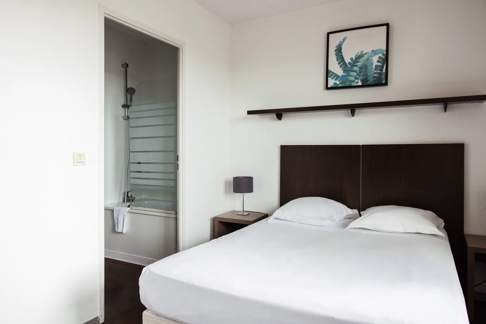 Modern bedroom with double bed with white linens, black headboard, adjoining bathroom with glass shower, minimalist décor and botanical artwork.